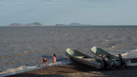 Two children play on a beach next to two beached metal boats. Mountains can be seen in the distance across the water.