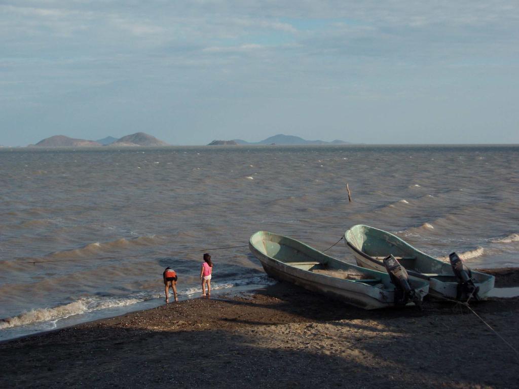 Two children play on a beach next to two beached metal boats. Mountains can be seen in the distance across the water.