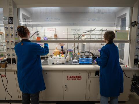 Two women in lab coats and glasses in a chemistry lab work under a fume hood.