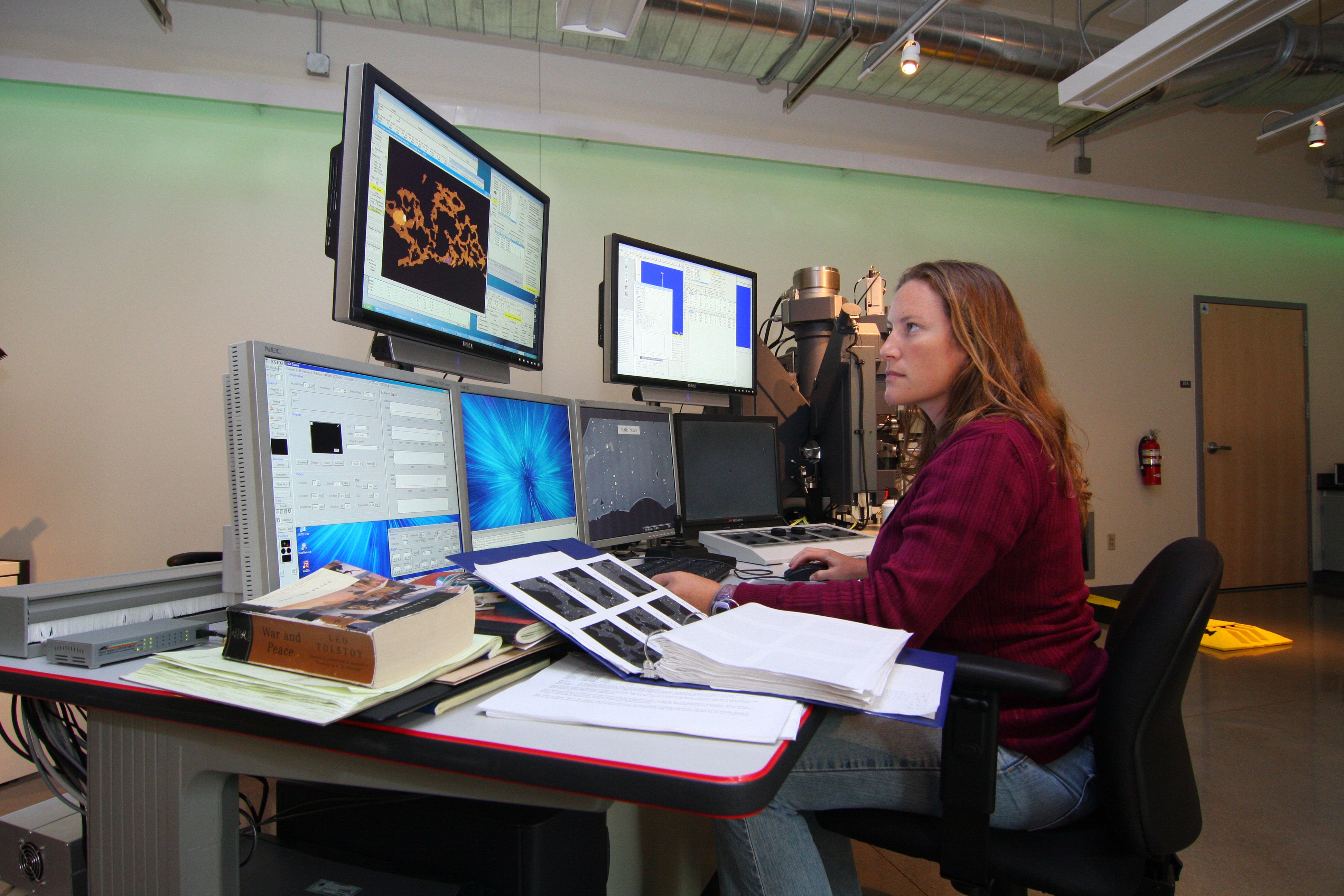 A woman sits at a computer station with multiple screens to compare data. There are open notebooks on the desk.