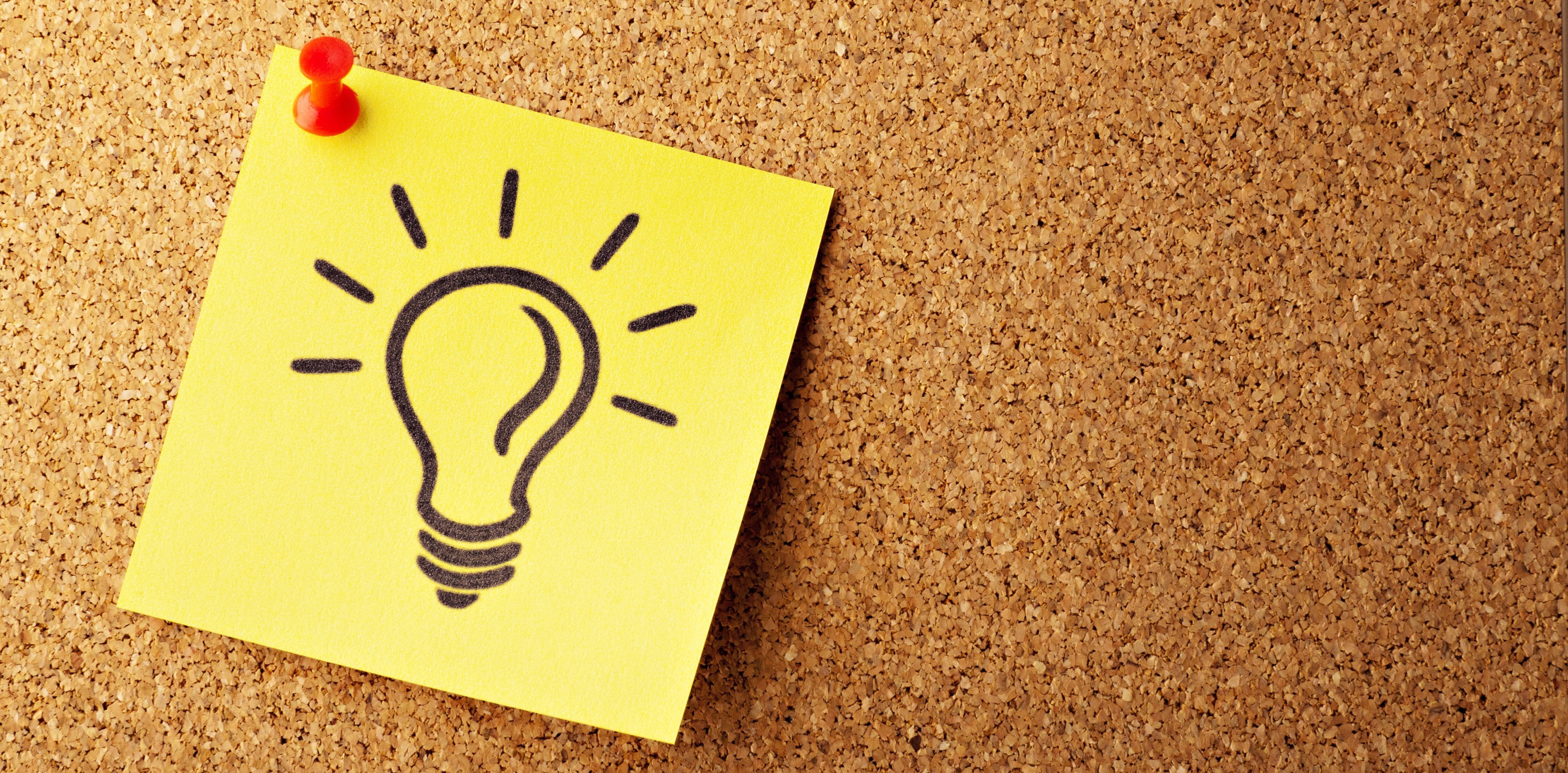 A yellow sticky note with a drawing of a light bulb is pinned to a cork board.