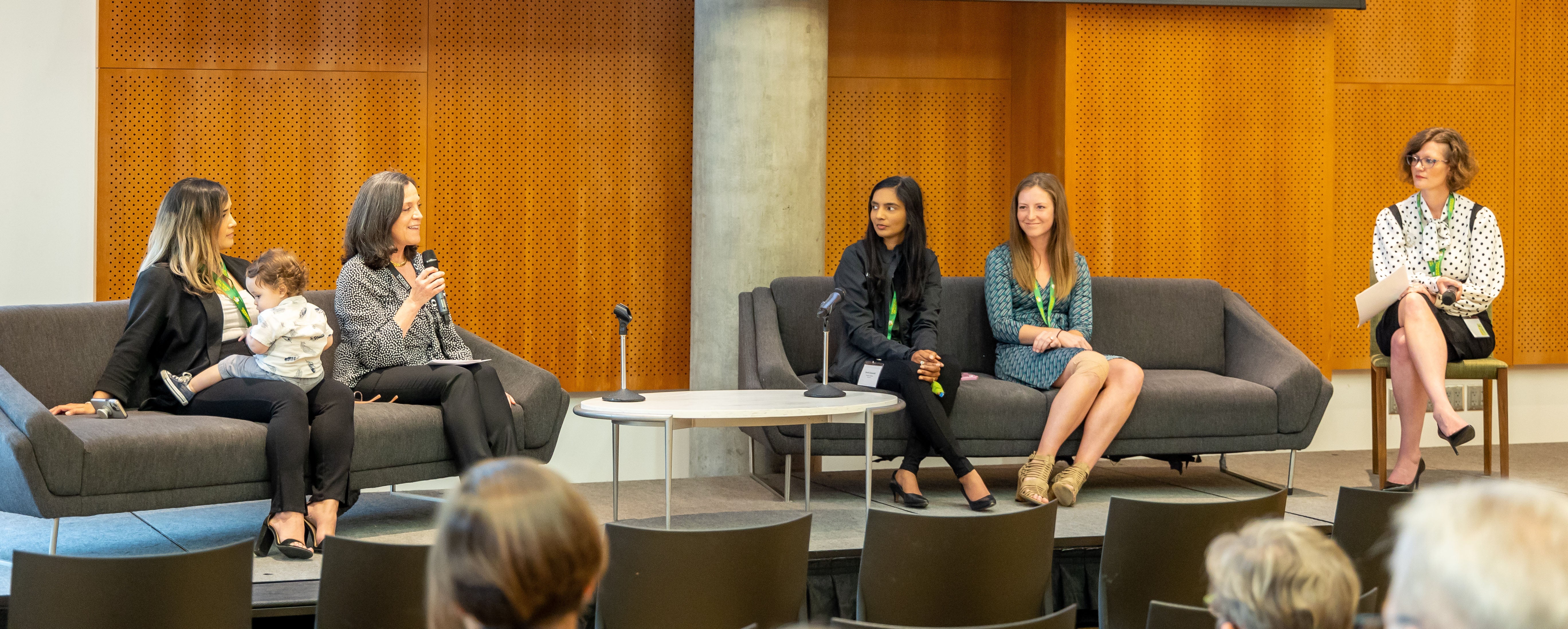 Four women participate in a panel for the women's innovation network seminar series. One woman has a small child on her lap and another woman is speaking into a microphone. A moderator is seated to the right of the panel. The audience is seen in the foreground of the image.