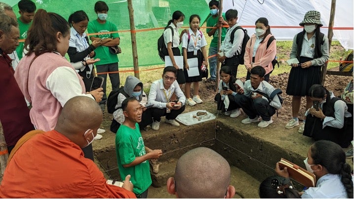 Schoolchildren stand around an archaeological dig site listening to an archaeologist.