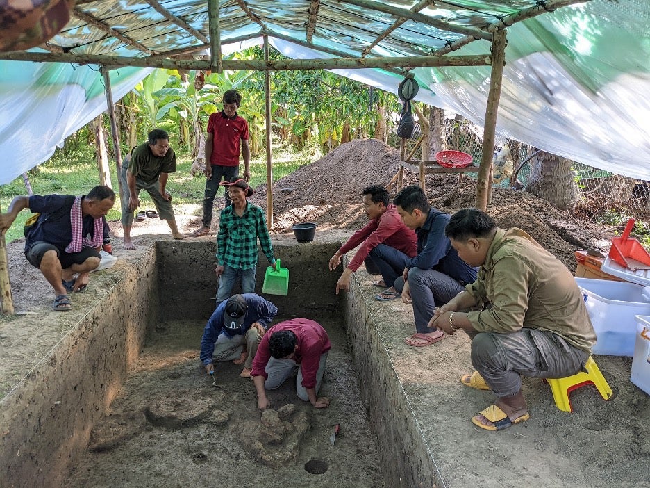 Under a plastic and wood pavilion, several people excavate at an archaeological site while others look on from above.
