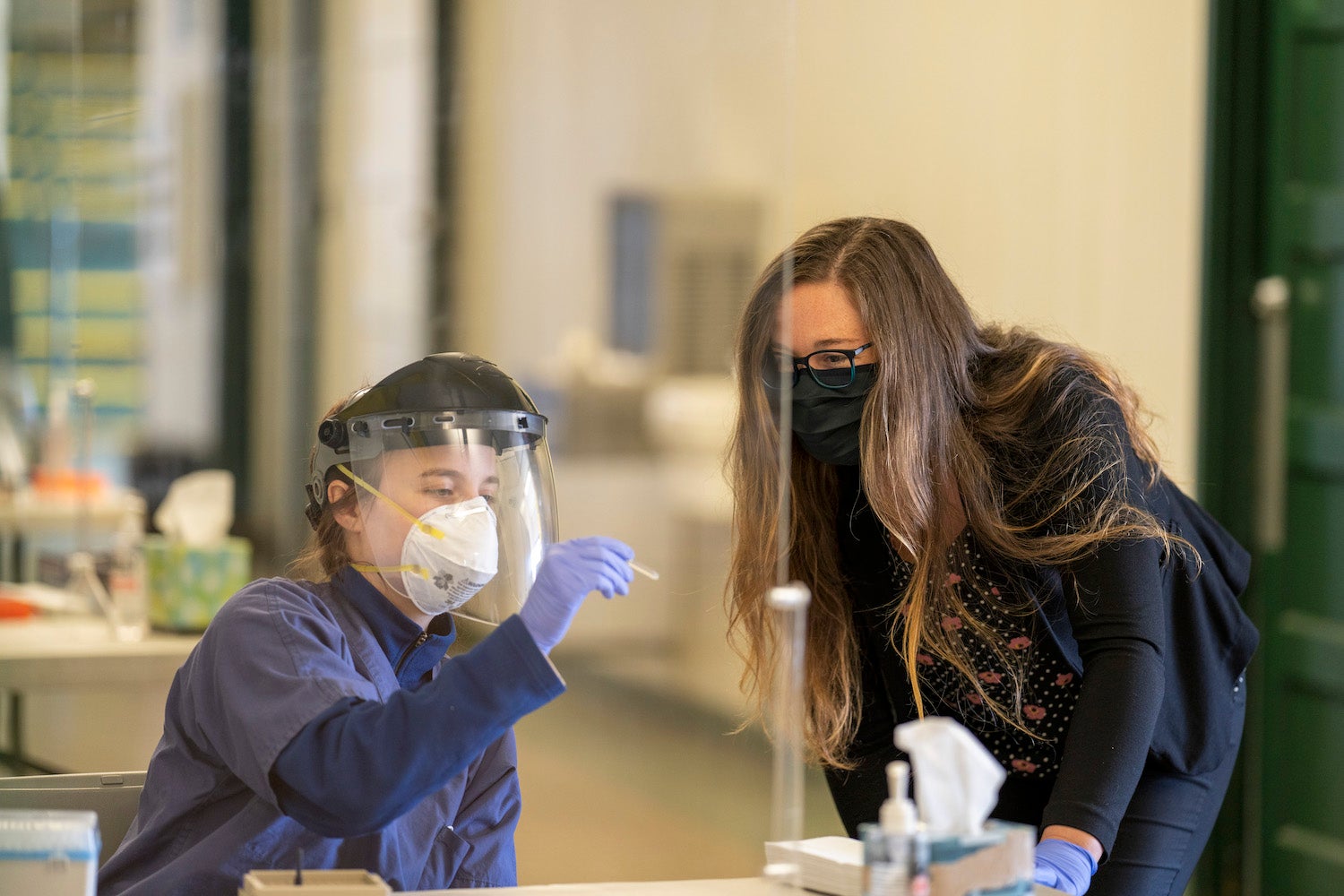 Two people wearing face masks and one also wearing a plastic face shield look at a COVID-19 sample in a plastic collection tube.