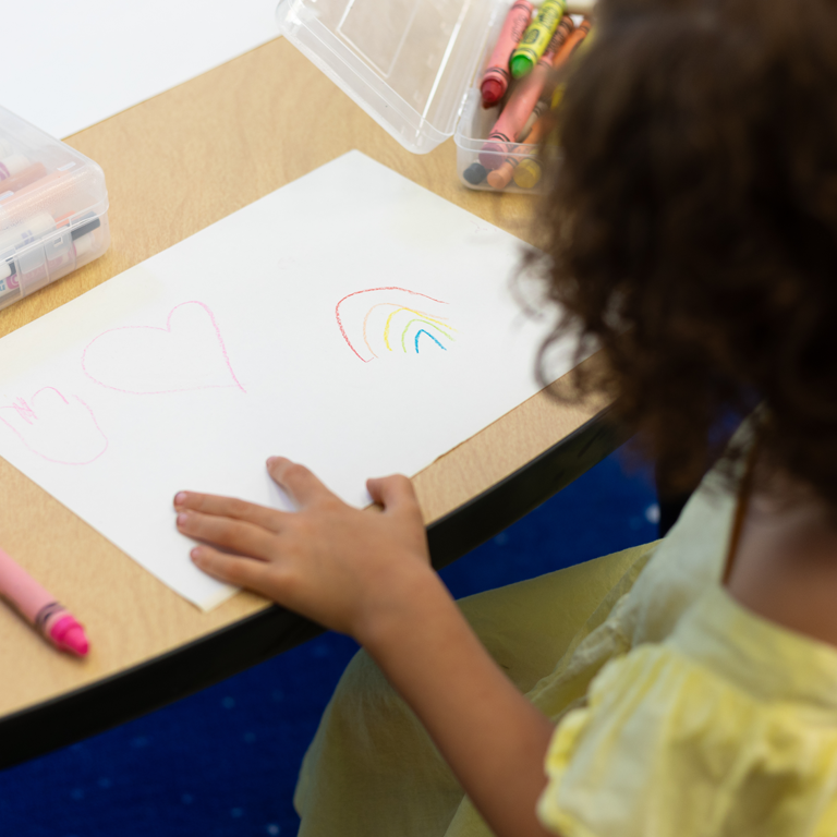A child looking at a crayon drawing of a rainbow and hearts.