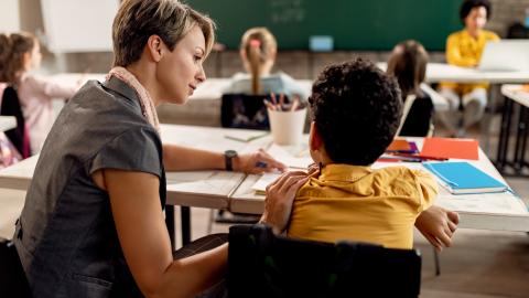 A female teacher sits at the side of young student with her hand on their shoulder while helping the child. The child is sitting at a desk in a classroom. 
