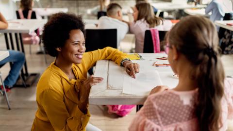 A female teacher smiles at a young female student while helping the student with a writing project. The teacher is squatting next to the desk the student sits at.