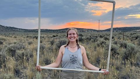 A portrait of Zoey Bailey, who is holding a square frame made of plastic PVC pipe, standing in front of a field of sagebrush at sunset.