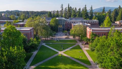 Arial picture of campus looking north from the Knight Library toward the Lillis Business Complex. In front of the building are a few students in a green, grass-covered quad. 