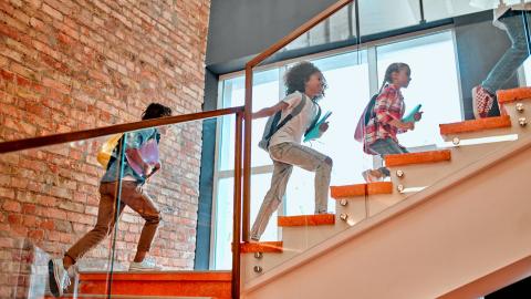 Four children wearing backpacks and carrying books run up a staircase in a school.