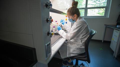 A woman wearing a lab coat and eye protection pipettes under a fume hood in a lab.