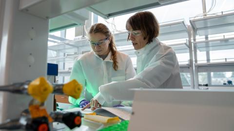 Two women wearing lab coats and eye protection do research in a laboratory.