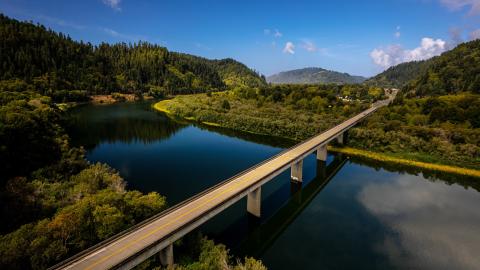 The Highway 101 bridge crosses over the Klamath River on a sunny day with the clouds reflected in the river's surface.