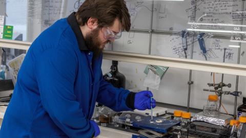 A scientist, wearing a lab coat and goggles, bends over a lab jack under the fume hood where he conducts his research. He is holding a syringe full of water over two cotton samples sitting at the lab jack.