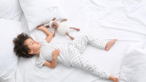 An African-American child sleeps in a bed with a sheep toy beside her.