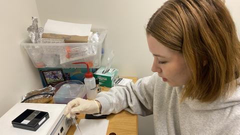 A young woman sits at a table wearing gloves and using a pipette. 