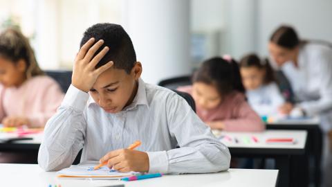 A young student rests his head on his hand while looking down at the paper on his desk.