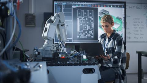 A researcher works on a laptop in an innovation space. Blueprints and formulas are displayed behind them.