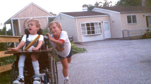 A young boy pushes another boy in a wheelchair in the driveway of a home.