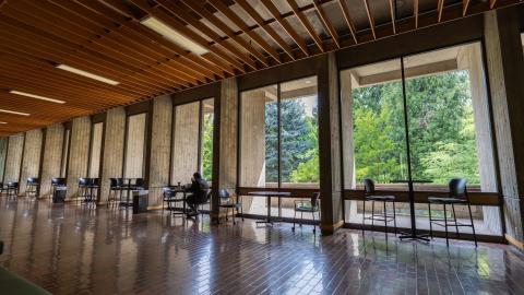 A person sits at a table in front of large floor-to-ceiling windows in McKenzie Hall on the UO campus.
