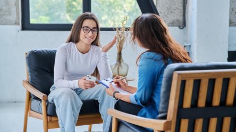 Two women face each other while sitting in arm chairs. One woman holds a cell phone and appears to be talking while the other listens while holding a clipboard.
