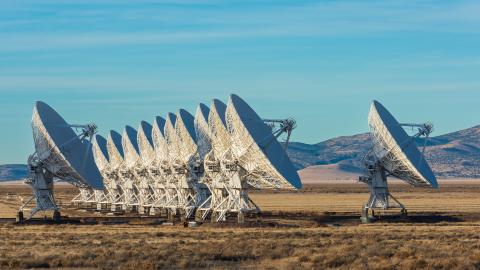 A long row of large white radio telescopes stands in an open desert plain under a blue sky.