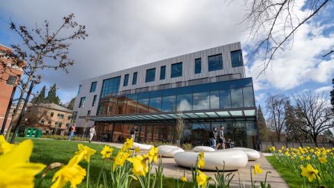 A modern multi-story glass and metal building on a college campus, with bright yellow flowers in the foreground.