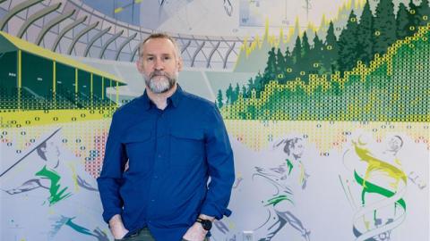 A portrait of a man, John Halliwill, standing in front of a mural of Hayward Field.