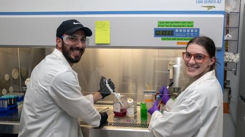 Two people in lab coats and safety glasses with pipettes in their hands stand in front of a fume hood in a laboratory smiling.