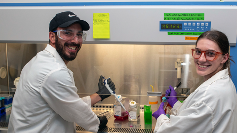 Two people in lab coats and safety glasses with pipettes in their hands stand in front of a fume hood in a laboratory smiling.