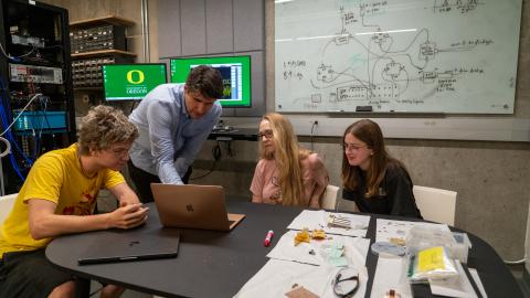A group of students look at a laptop as a faculty member points to something on the screen.