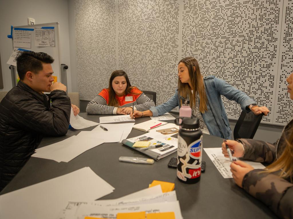 Four people gathered around a table scattered with papers brainstorm together.