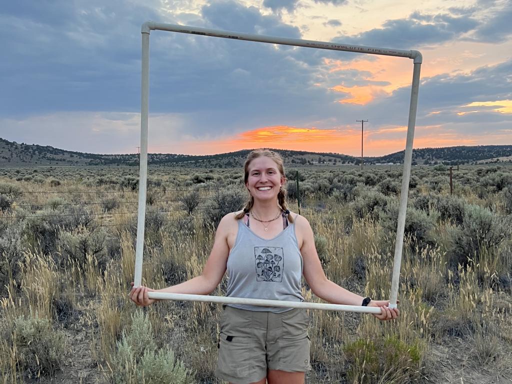 A portrait of Zoey Bailey, who is holding a square frame made of plastic PVC pipe, standing in front of a field of sagebrush at sunset.
