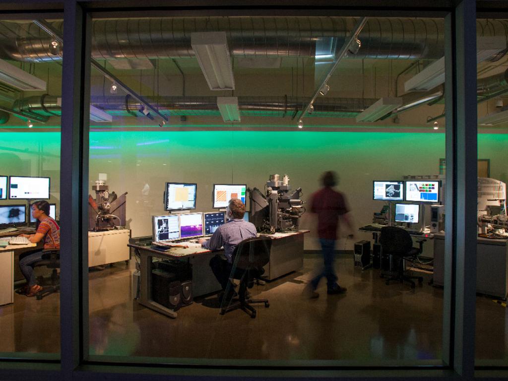 Three computer stations with people sitting at them or walking past them seen through glass doors.