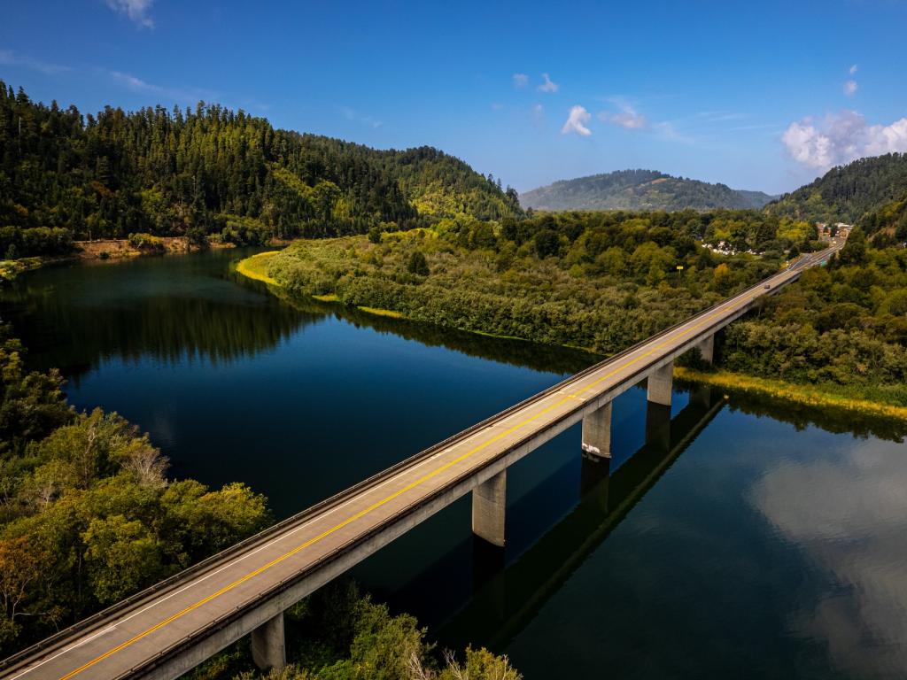 The Highway 101 bridge crosses over the Klamath River on a sunny day with the clouds reflected in the river's surface.
