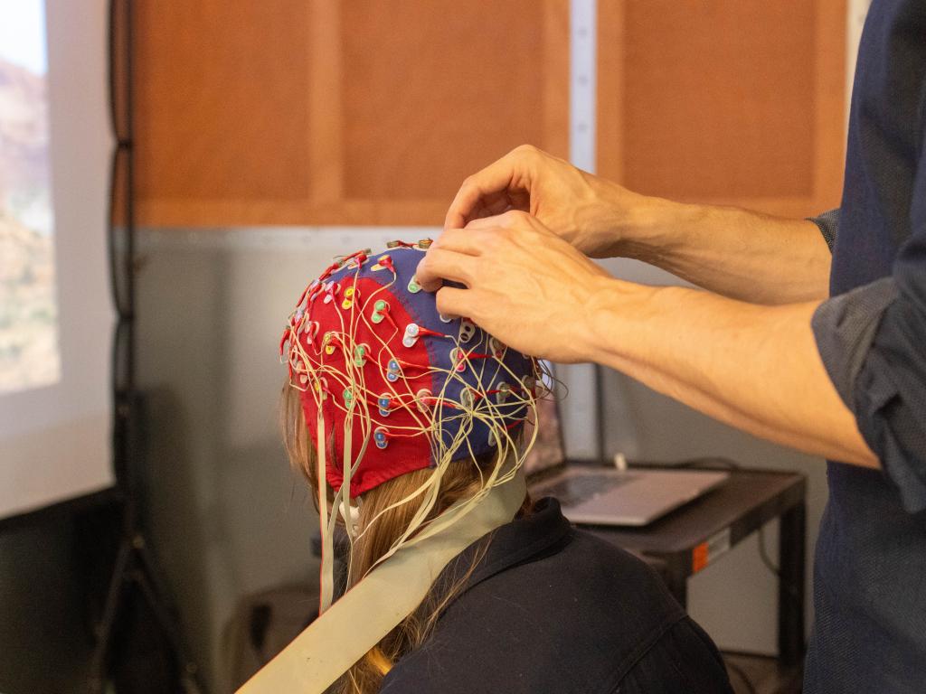 Research participant sits in a chair and has an electroencephalogram (EEG) cap placed on their head.  