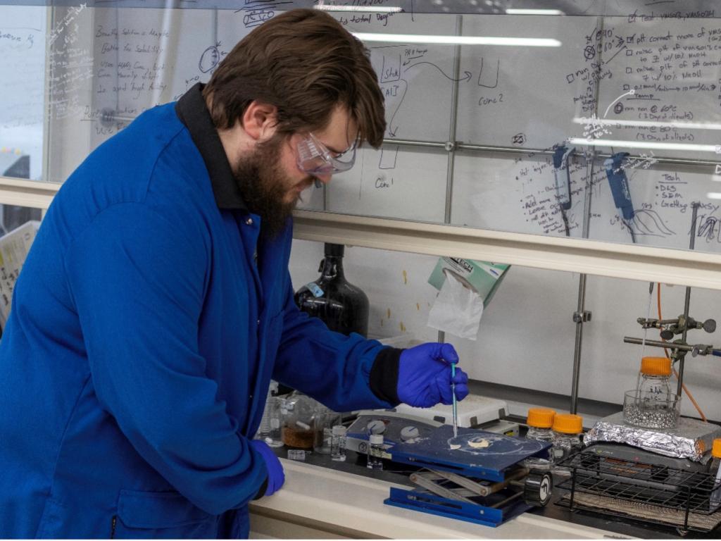 A scientist, wearing a lab coat and goggles, bends over a lab jack under the fume hood where he conducts his research. He is holding a syringe full of water over two cotton samples sitting at the lab jack.