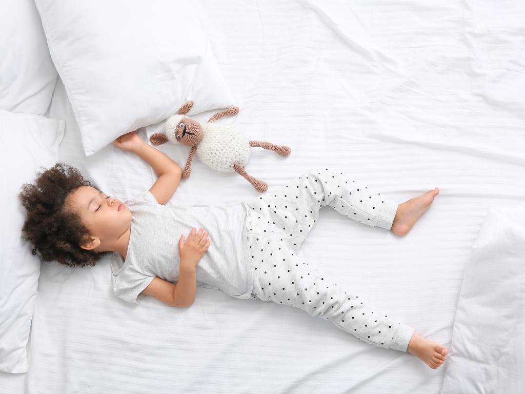 An African-American child sleeps in a bed with a sheep toy beside her.