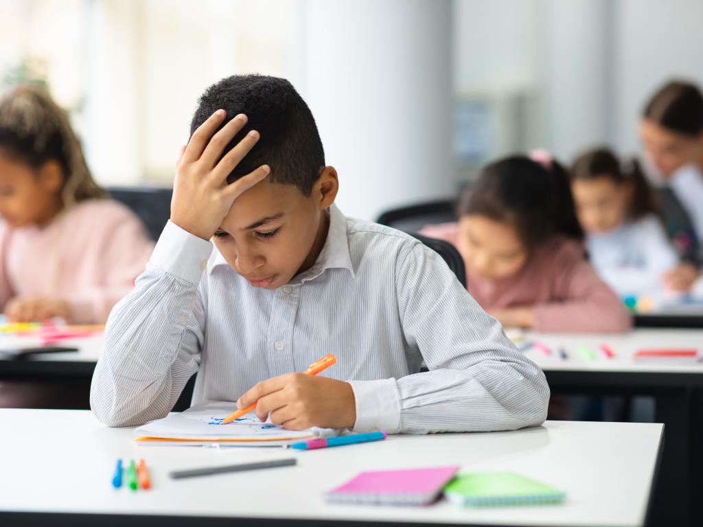 A young student rests his head on his hand while looking down at the paper on his desk.