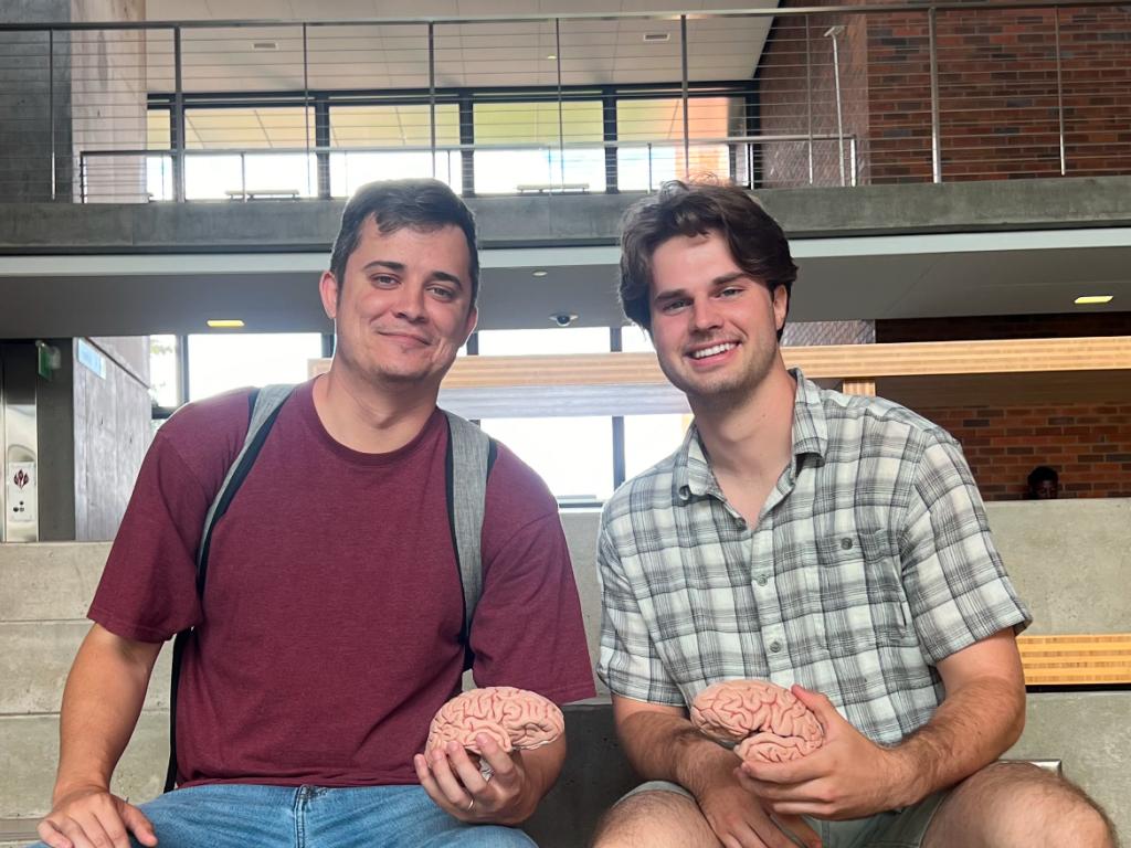 Two students sit on stairs in the Lewis Integrative Science Building, holding models of brains.