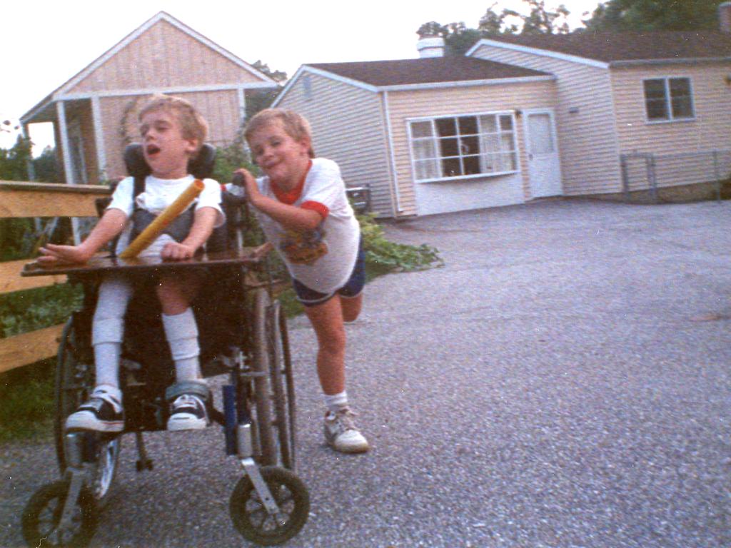 A young boy pushes another boy in a wheelchair in the driveway of a home.