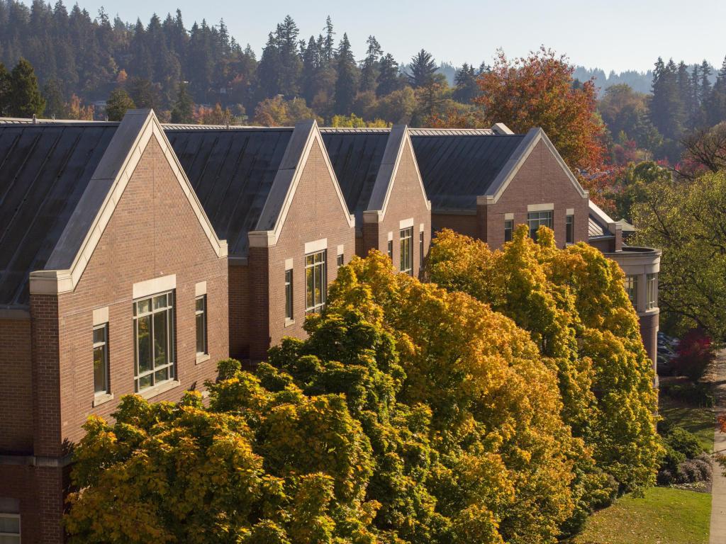 A red brick building lined on the front with trees with green, yellow, and orange leaves.