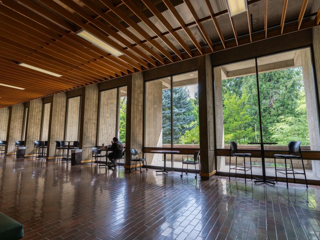 A person sits at a table in front of large floor-to-ceiling windows in McKenzie Hall on the UO campus.