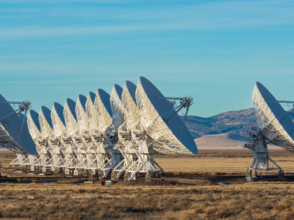 A long row of large white radio telescopes stands in an open desert plain under a blue sky.