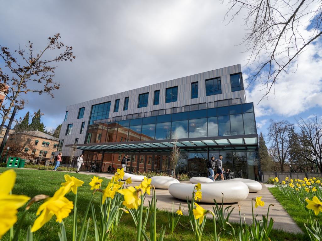 A modern multi-story glass and metal building on a college campus, with bright yellow flowers in the foreground.