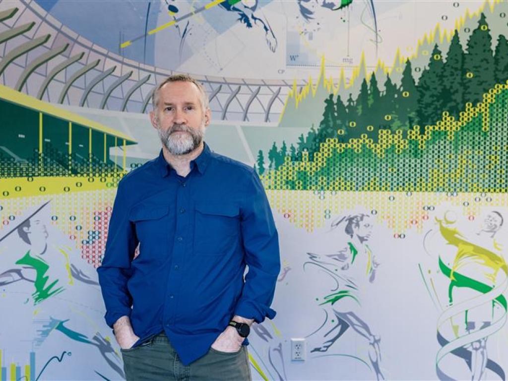 A portrait of a man, John Halliwill, standing in front of a mural of Hayward Field.