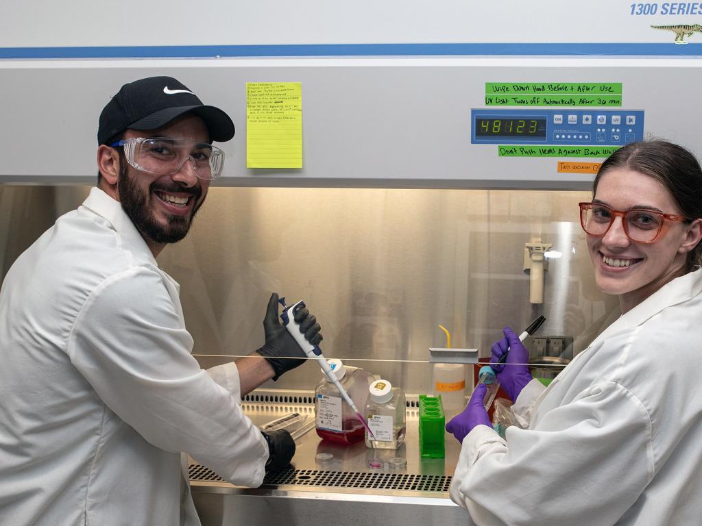 Two people in lab coats and safety glasses with pipettes in their hands stand in front of a fume hood in a laboratory smiling.
