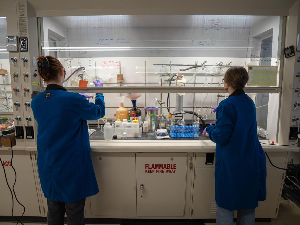 Two women in lab coats and glasses in a chemistry lab work under a fume hood.