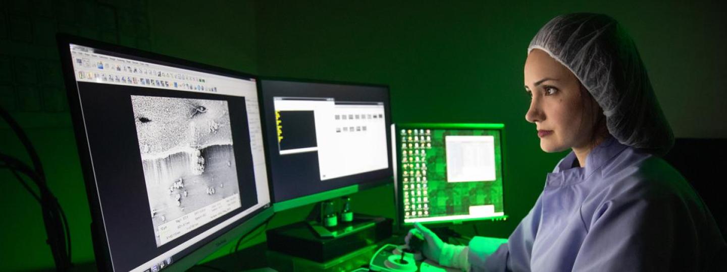 A woman wearing a hairnet, lab gown, and gloves uses a joystick to adjust the image on a computer monitor.